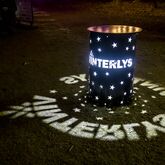En lysinstallation fra Vinterlys Festivalen i Skjern. En tønde med laserskårne stjerner og teksten "Vinterlys" projicerer lyset ned på jorden og skaber en stemningsfuld effekt i mørket.
